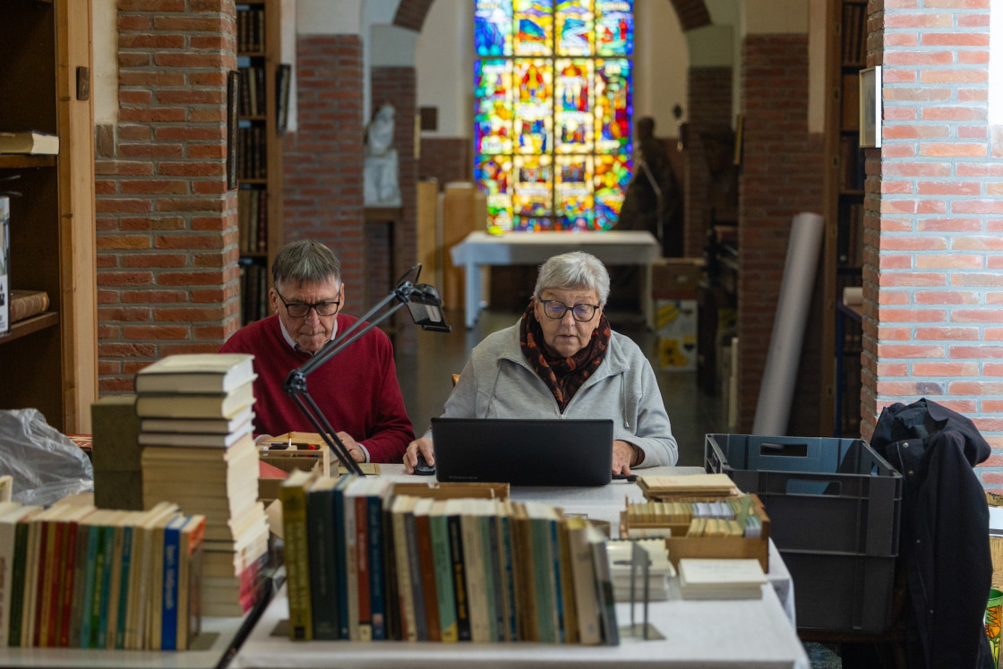 Twee vrijwilligers aan het werk in de bibliotheek van de abdij van Postel. Door het glasraam op de achtergrond valt kleurig licht naar binnen.