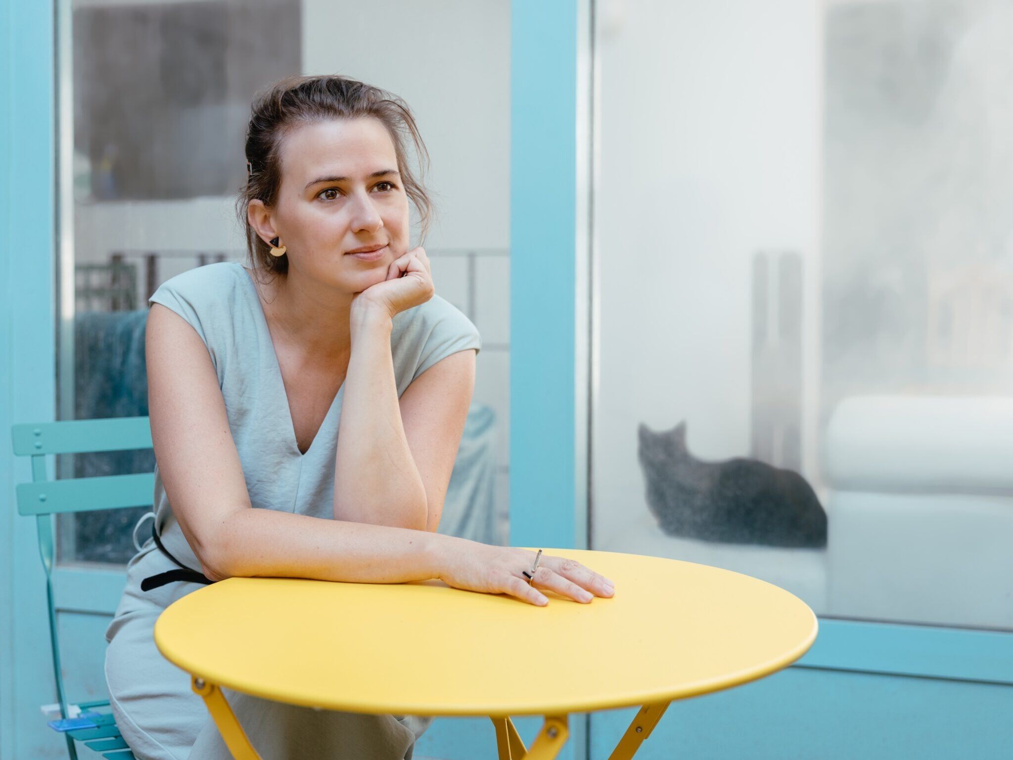 Maud Vanhauwaert dromerig aan een terrastafeltje. Kat op de vensterbank.