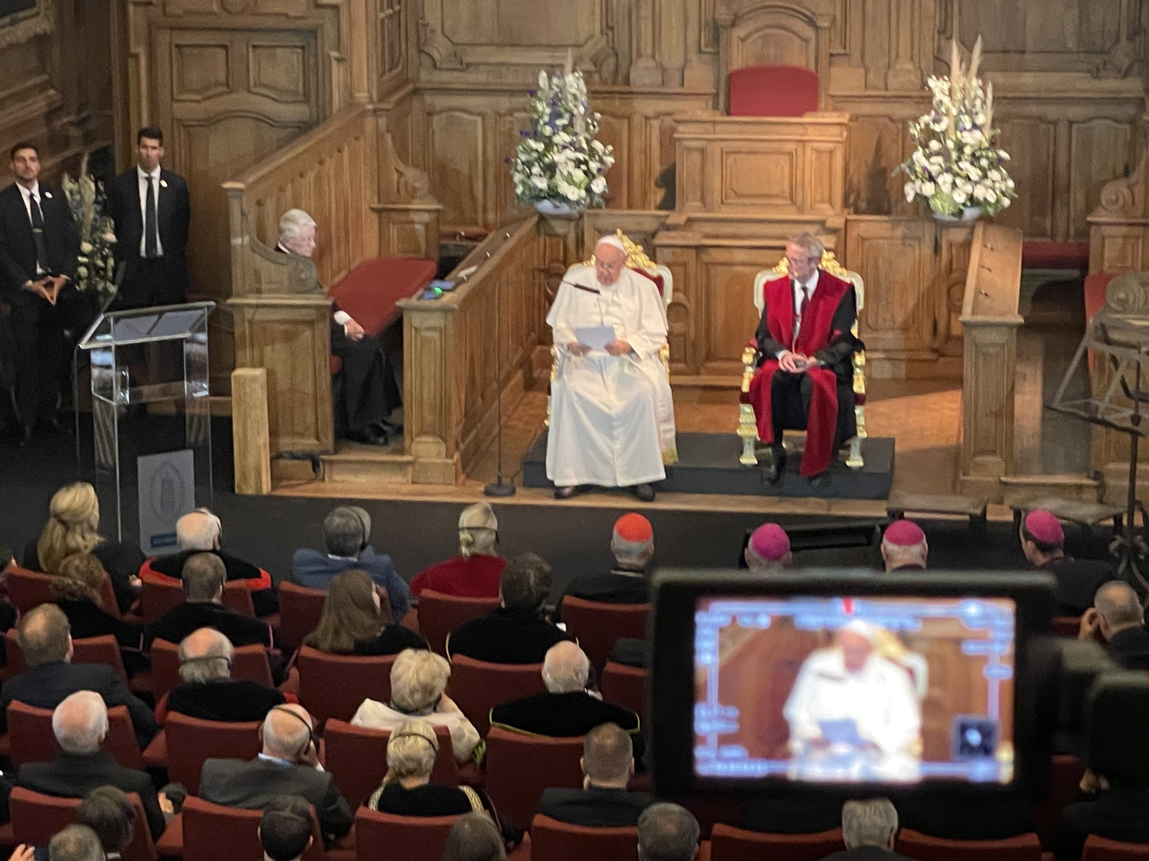 Paus Franciscus en rector Luc Sels zitten vooraan in de Promotiezaal van de KU Leuven. Op de voorgrond een camera met hetzelfde beeld.