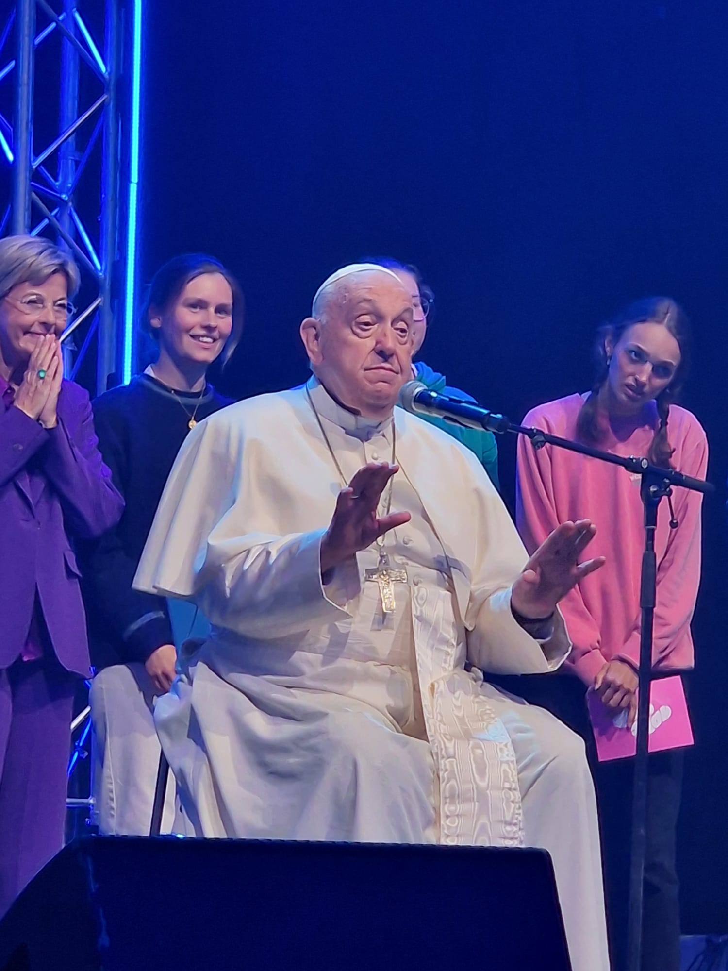 Paus Franciscus spreekt jongeren toe op Hope Happening. Achter hem staat Ingeborg in paars broekpak en de handen blij verrast tegen elkaar voor de mond geslagen.