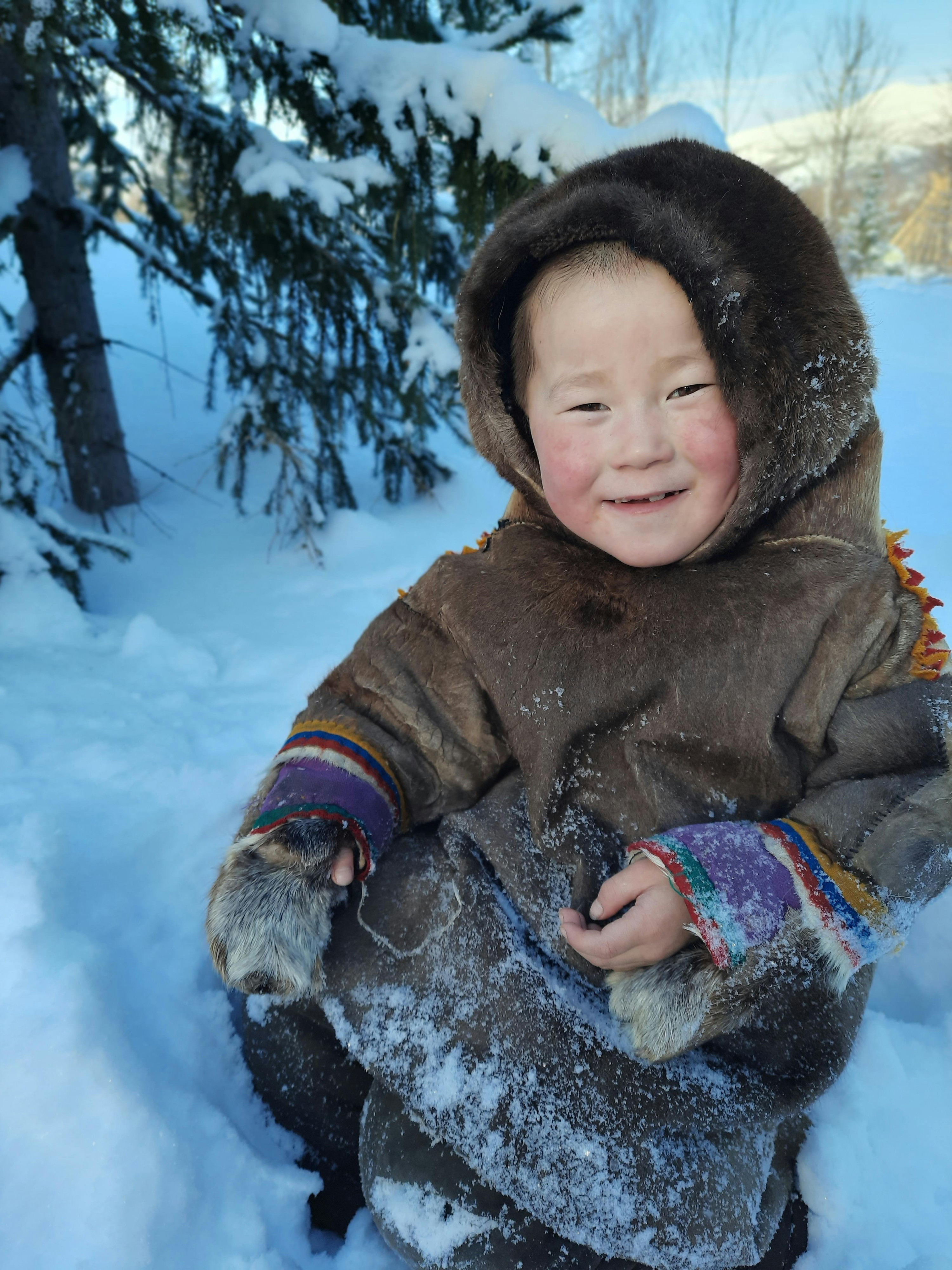 Een Inuit kleuter zit in de sneeuw en kijkt de camera lachend aan.