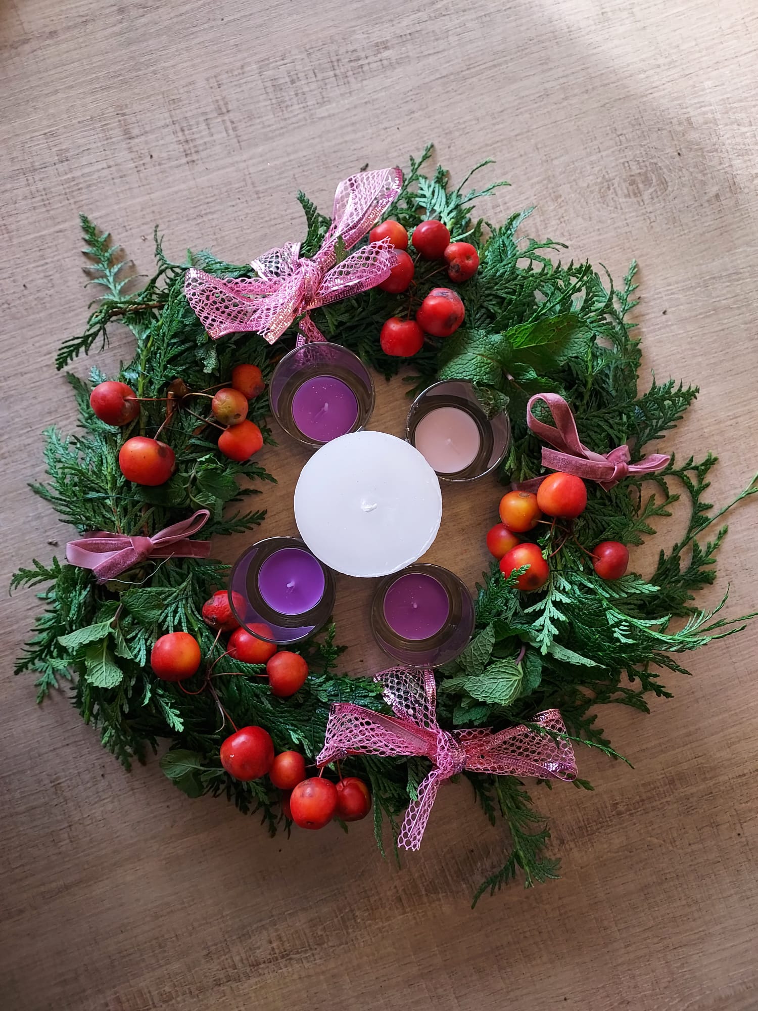 Een adventskrans met drie paarse en één roze theelichtje rondom een centrale witte kaars.