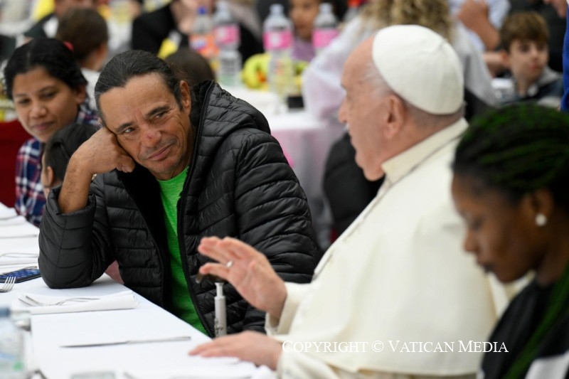 Paus Franciscus praat levendig met een man naast hem aan de tafel, die luistert met een geamuseerde lach op het gezicht.